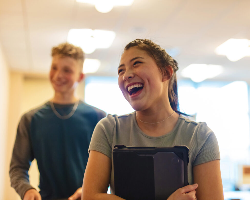 A young woman laughs while holding a tablet. A young man stands behind her smiling.