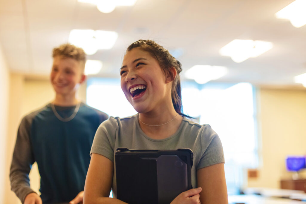 A young woman laughs while holding a tablet. A young man stands behind her smiling.